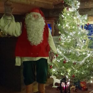 Joulupukki heiluttaa kameralle hirsituvassa. Joulupukin vieressä kauniisti koristeltu joulukuusi, jonka alla on lyhty, pieni puinen keinuhevonen ja kuorma-auto lelu.